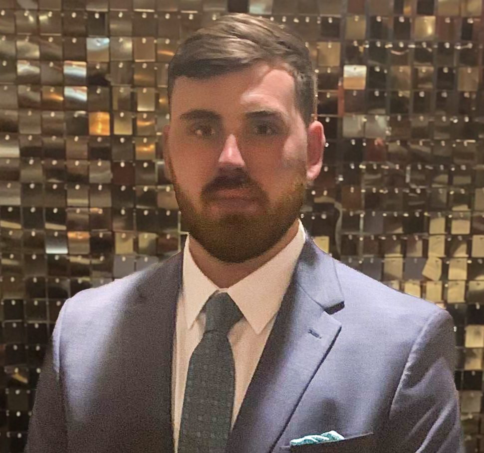 A man with short dark hair and a beard is wearing a light gray suit with a light blue tie and pocket square. He is standing in front of a shiny, metallic background composed of small, square tiles, possibly promoting life insurance services in Columbus, GA.