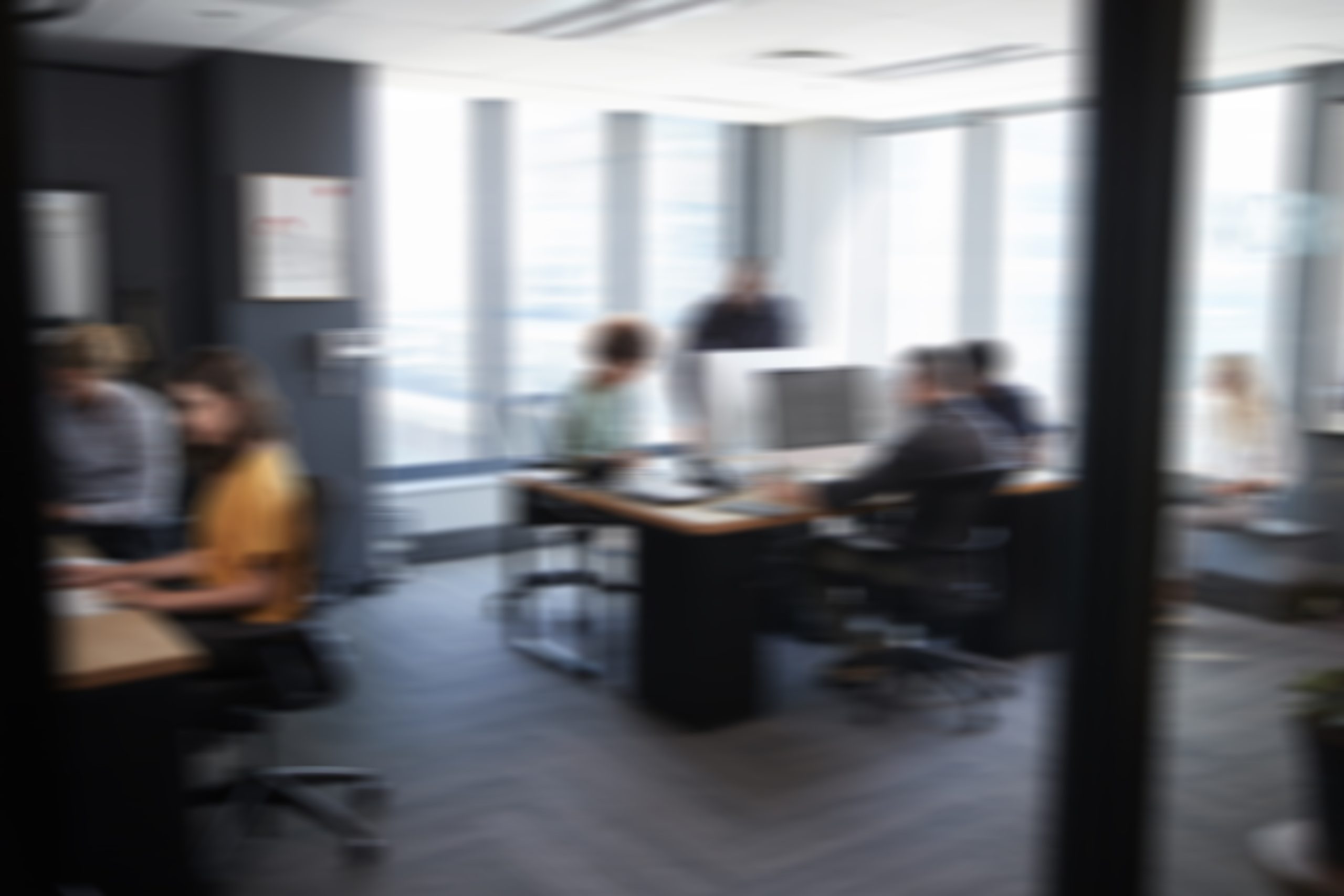 A blurred image of a modern office setting in Columbus, GA, with several people working at desks and standing near a large window. The room has natural light coming in through the windows, and the workspace is furnished with computers, office chairs, and various office supplies related to life insurance.